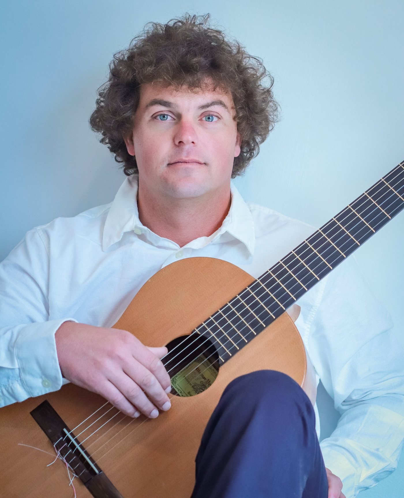 Richard Knowles seated with a classical guitar, wearing a white shirt against a soft blue wall